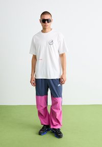 Man wearing oversized white T-shirt with graphic, color-block navy and pink Nike pants, black sneakers, and large black sunglasses, standing on green floor.