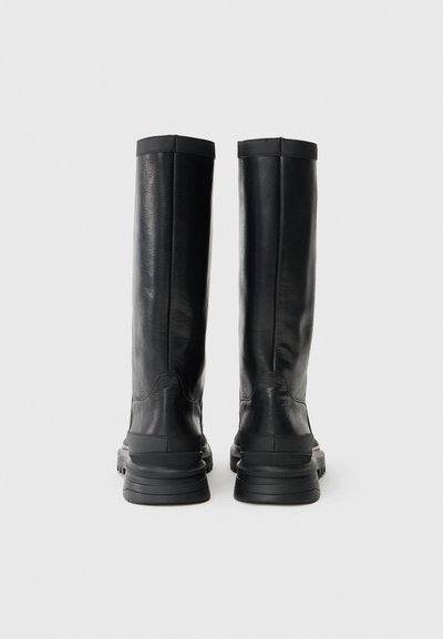 Svarta läderknähöga stövlar med en strukturerad yta, platta sulor och en stilren design. Utrustade med en enkel stickad kant.