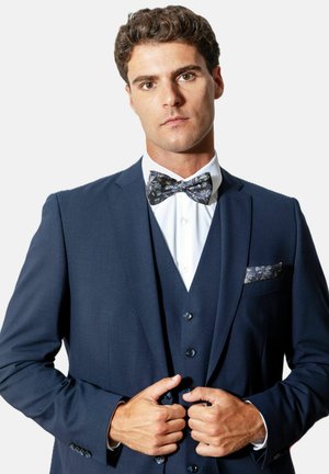 Homme boutonnant un costume trois pièces bleu marine avec un nœud papillon fleuri et une pochette assortie, chemise blanche, fond neutre.