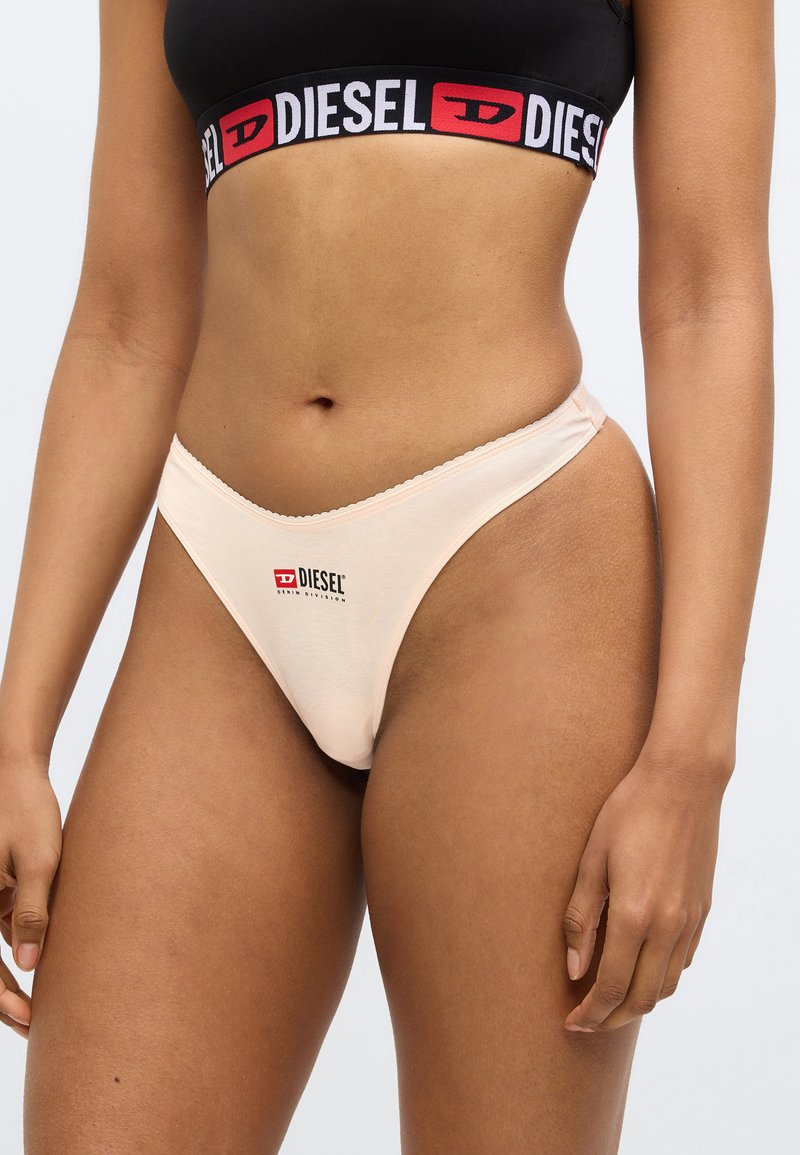 Peach-colored thong with a smooth texture, thin straps, and a DIESEL logo in red and black on the front.
