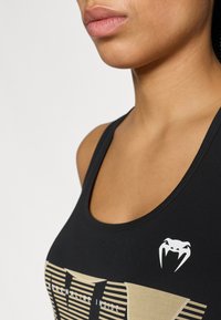 Musta hihaton paita, jossa on kultainen raidallinen kuvio ja painettu teksti "ADRENALIINI SISÄLLÄ." Vasemmalla puolella valkoinen logo.