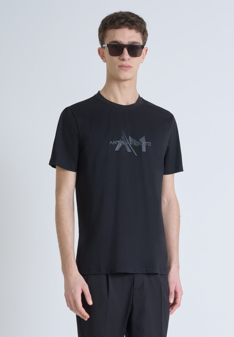 Camiseta negra de cuello redondo hecha de algodón suave, con un discreto logo tonal "AM" en el frente. El modelo lleva gafas de sol negras.