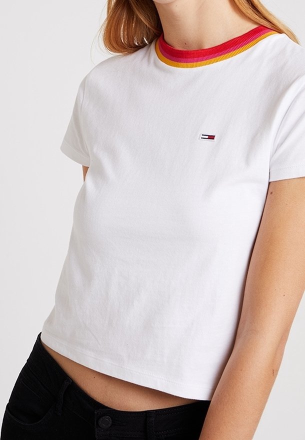 Mujer vistiendo una camiseta corta de manga corta blanca con cuello a rayas rojas y amarillas y un pequeño logo en el pecho, combinada con pantalones negros.