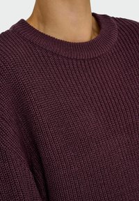 Lähikuva henkilöstä, joka on pukeutunut tumman violettiin, paksusti neulottuun villapaitaan, jossa on ribbi-kaulus.
