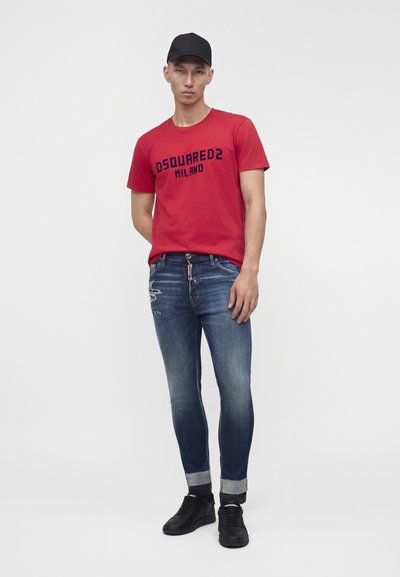 Νέος άνδρας που φοράει ένα κόκκινο t-shirt DSQUARED2 Milano, κατεβασμένο μπλε τζιν με φθορές, μαύρα αθλητικά παπούτσια και μαύρο καπέλο μπέιζμπολ, στέκεται με το ένα χέρι πίσω.