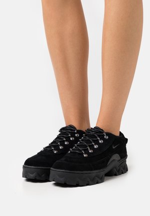 Czarne buty trekkingowe Nike z grubą, wytrzymałą podeszwą i metalowymi przelotkami, noszone na gołych nogach na białym tle.