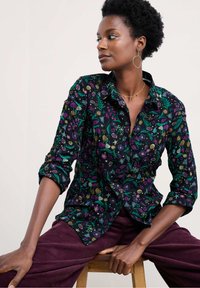 Camicia a maniche lunghe con bottoni, nera, decorata con motivi floreali in viola, verde e oro. Maniche arrotolate ai polsi. Abbinata a pantaloni in velluto a coste.