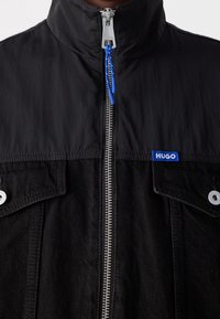 Giacca nera con colletto alto, zip argentata con un a strappo blu, due tasche a patta con bottoni argentati e una patch blu con il logo HUGO.