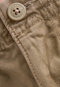 Beige tyg med en slät yta, med en knapp med "SUPERDRY" präglad, synlig sömnad och tygveck.