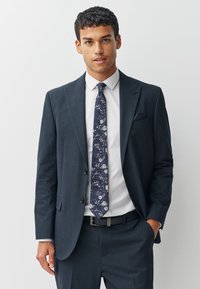 Costume texturé bleu marine avec une chemise blanche et une cravate à motifs floraux. Présente des revers traditionnels et un mouchoir de poche dans la poche poitrine.