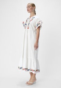 Abito lungo in cotone bianco con ricami floreali in blu, rosso e verde. Caratterizzato da maniche corte a volant e una orlo a strati. Si apre con una cerniera frontale.