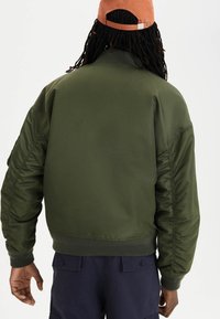 Olivově zelená bomber bunda s ribbovaným lemem a manžetami, vybavená strukturovanými rameny a hladkým, lesklým povrchem. Žádné viditelné vzory.