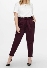 Kobieta w białej koszuli z krótkim rękawem, włożonej w wysokie bordowe spodnie typu paperbag z przewiązanym paskiem i czarnych butach na wysokim obcasie.
