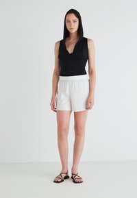 Black sleeveless top with a hood, paired with white shorts featuring eyelet embroidery along the hem. Model stands in a neutral backdrop.