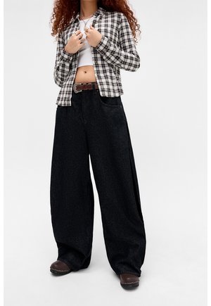 Persona che indossa jeans neri a gamba larga, una cintura marrone, scarpe marroni, un top corto bianco e una camicia a quadri bianca e nera con bottoni.