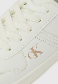 Λευκό δερμάτινο αθλητικό παπούτσι με υφή στην επιφάνεια, που διαθέτει κρεμ κορδόνια και ροζ χρυσό λογότυπο "ck" στη πλευρά. Απλός σχεδιασμός και χαμηλό προφίλ.