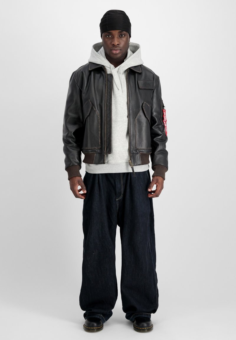 Brun läderbomberjacka med ribbade manschetter, grå huvtröja och lösa mörkblå denimbyxor. Svarta skor fulländar outfiten.