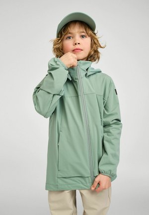 Child with light brown hair wearing a green cap and green jacket, zipping up the jacket against a plain light background.
