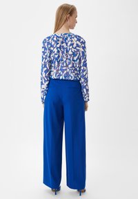 Blaue hoch taillierte Hosen mit weitem Bein, kombiniert mit einer transparenten Bluse mit langen Ärmeln und einem blau-weißen abstrakten Muster.