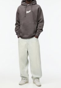 Sudadera gris con capucha y bolsillo frontal, con un logo blanco. Jeans de pierna ancha en verde claro. Zapatillas blancas con un diseño texturizado.