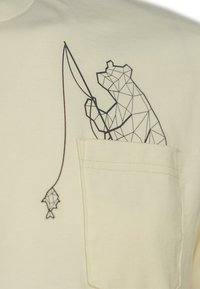 Camiseta de manga corta en color crema con un gráfico geométrico de un oso y un pez en negro. Detalle de bolsillo en el lado izquierdo. Textura de tela suave.