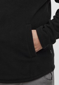 Svart fleece genser med teksturert overflate. Har en sidelomme og elastiske mansjetter. Diskret merkevare ved kantene.