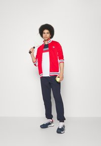 Rød hettejakke med hvite og marineblå striper, hvit t-skjorte med røde striper, marineblå bukser og marineblå joggesko. Holder en tennisracket og tennisball.