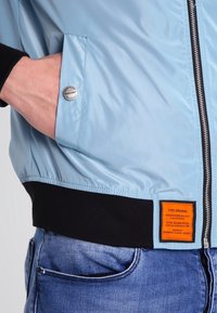 Světle modrá bomber bunda s černými žebrovanými manžetami a lemem. Má stříbrné patentní knoflíky a oranžovou etiketu se specifikacemi.
