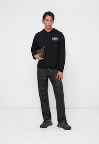 Hoodie noir avec poche avant et graphique coloré, associé à un pantalon en cuir noir. Tenant un petit sac noir texturé.