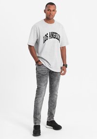 Harmaan puuvillapaita, jossa lukee "LOS ANGELES" mustalla, yhdistettynä haalistuneisiin harmaisiin farkkuihin ja mustiin lenkkareihin, seisoo valkoista taustaa vasten.