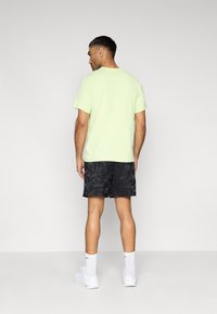 Ανοιχτοπράσινο πουκάμισο με κοντό μανίκι συνδυασμένο με μαύρα σορτς καμουφλάζ. Η εμφάνιση έχει άνετη εφαρμογή και ολοκληρώνεται με λευκά αθλητικά παπούτσια.