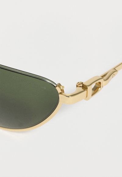 Γυαλιά ηλίου με χρυσό μεταλλικό σκελετό και σκουρό πράσινους φακούς, που δείχνουν λεπτομερή σχεδιασμό μεντεσέδων και βραχίονα σε λευκό φόντο.