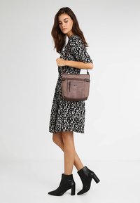 Braune Leder-Schultertasche mit einer vorderen Reißverschlusstasche, verstellbarem Gurt, eleganter Form und glatter Textur, ergänzt durch ein stilvolles schwarzes Kleid.