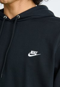 Črna kapuca z vrvicami, ki ima na prsih belo Nike logotip. Gladko teksturo, sproščen kroj in rebraste manšete.
