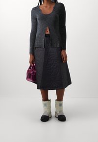 Maglia nera a maniche lunghe con una texture a coste e uno spacco frontale, abbinata a una gonna trapuntata scura, stivali bianchi e neri e una borsa lucida rosa.
