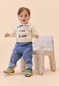 Kleinkind steht neben einem gepunkteten modernen Stuhl, trägt einen weißen Pullover mit buntem Text, blaue Jeans und mehrfarbige Schuhe.