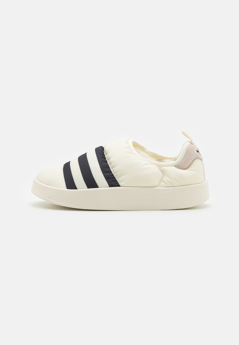 Baskets sans lacets couleur crème avec des rayures bleu marine foncé, semelle en caoutchouc, tige rembourrée et un petit onglet beige au talon avec le logo Adidas.