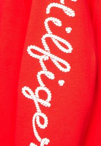 Czerwona bluza z białymi, sznurkowymi literami na przodzie. Materiał wydaje się miękki o gładkiej fakturze. Projekt podkreśla wyraźny kontrast kolorów.