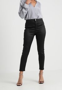 Svarta högmidjade byxor med en figurnära siluett, tillverkade av ett slätt tyg. Kombinerat med en ljusgrå v-ringad blus och remsandaler.