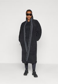 Zwarte fleece lange jas met een rits aan de voorkant, zijzakken en een zachte textuur, gecombineerd met een zwarte outfit en een zonnebril.