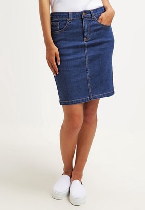 Vrouw die een blauwe spijkerrok tot op de knie draagt, witte instapschoenen, met één hand in de zak en de andere langs haar zijde.