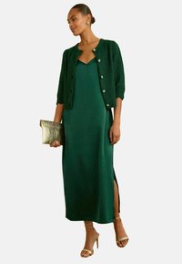 Rochie maxi din satin verde cu o fendue până la coapsă, asortată cu un cardigan tricotat de culoare verde închis și sandale cu toc aurii cu curele, ținând o poșetă aurie.