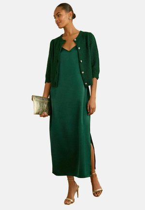 Green satin maxi dress with a thigh-high slit, paired with a knitted dark green cardigan and gold strappy heels, holding a gold clutch bag.