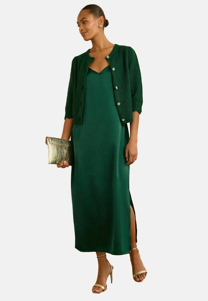 Rochie maxi din satin verde cu o fendue până la coapsă, asortată cu un cardigan tricotat de culoare verde închis și sandale cu toc aurii cu curele, ținând o poșetă aurie.