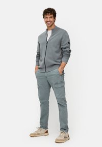 Lichtgrijze zip-up trui met geribbelde accenten, gecombineerd met lichtgrijze cargobroek met zijzakken en beige sneakers met bruine accenten.