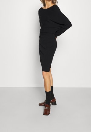 Kobieta ubrana w czarną, dopasowaną sukienkę z długim rękawem, czarne skarpetki i brązowe mokasyny na obcasie, stojąca na jednolitym białym tle.