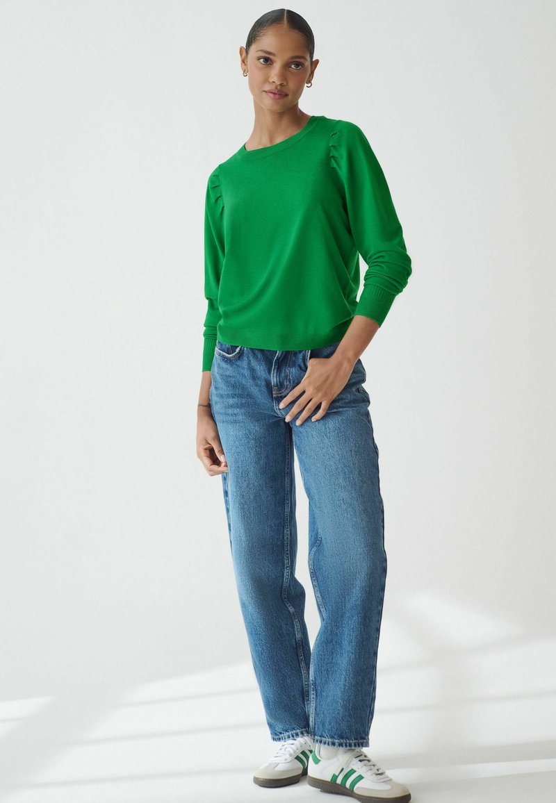 Grøn strikket sweater med lange ærmer, ribbede manchetter og diskrete sideløsninger, parret med lyseblå højttaljerede jeans og hvide sneakers.