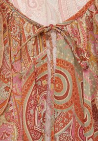 Multikleurige stof met complexe paisley- en bloemenpatronen in tinten rood, oranje, roze en beige; knoopdetail bij de hals.