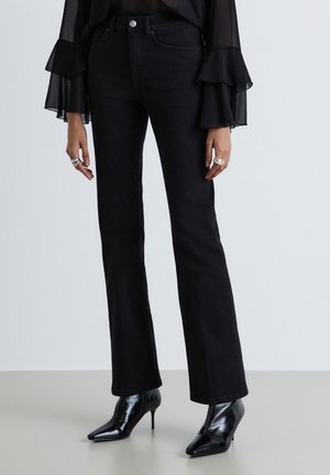 Femme portant un jean flare noir, un chemisier noir transparent à manches volantées, des bagues argentées et des bottines noires pointues à talons hauts.