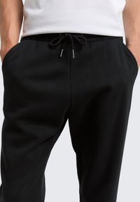 Pantalones de chándal negros con cintura elástica y cordón. Cuenta con bolsillos laterales y un tejido suave y texturizado. Diseño minimalista.